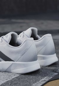 Paire de chaussures de course athlétiques gris clair avec l’étiquette « DURAMORC2 » sur le côté, posées sur une surface extérieure texturée.