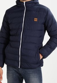 Marinblå puffjacka med dragkedja fram, horisontellt quiltat mönster, huva och logopatch i brunt läder på vänster bröst.