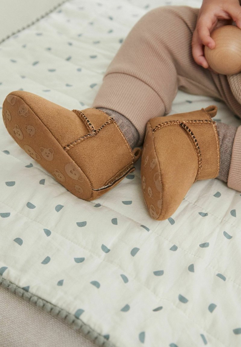 Bébé portant des chaussures en daim marron à semelle souple avec des motifs d'ours, pantalon beige, tenant une balle en bois en étant assis sur une courtepointe à motifs.