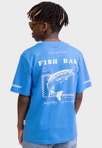 Camiseta de algodón azul con un estampado gráfico de un pez, detalles de cuadros y texto en la parte posterior. Mangas cortas y un diseño de ajuste relajado.
