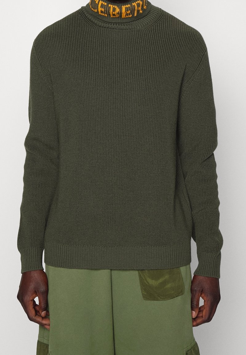 Pull en maille vert olive foncé avec poignets et ourlet côtelés, porté avec un pantalon vert doté d'une poche sur la cuisse.