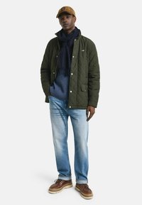 Quilted grüne Jacke mit einem marineblauen Schal, hellblauem Pullover, ausgebleichten blauen Jeans und braunen Schuhen. Die Mütze ist kariert in Gelb- und Brauntönen.