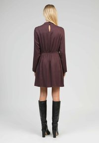 Femme aux longs cheveux blonds portant une robe bordeaux jusqu'aux genoux et des bottes noires à talons hauts jusqu'aux genoux, se tenant de dos contre un fond uni.