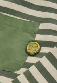 Groen en crème gestreept stof met een effen groene zak en een ronde gele en grijze badge die over de zak heen ligt.