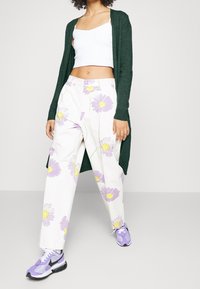 Valkoiset housut, joissa violetteja ja keltaisia kukkakuoseja, yhdistetty vihreään neuletakkiin ja lilaan tennareihin. Sileä materiaali, rento istuvuus.