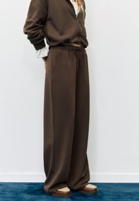 Smeđa dukserica na patentni zatvarač s opuštenim krojem i prugastim rukavima; uparena s opuštenim smeđim trenirkama i cipelama s kremastim detaljima.