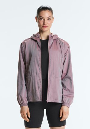 Kobieta o ciemnych włosach związanych w kucyk, ubrana w jasnofioletową bluzę z kapturem zapinaną na zamek oraz czarne sportowe spodenki i top, stojąca na jednolitym tle.