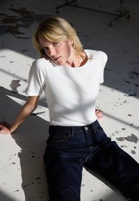 Femme blonde en haut blanc à manches courtes et jean foncé, assise sur un sol usé éclaboussé de peinture, avec la lumière du soleil projetant des ombres autour d'elle.