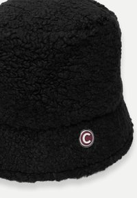 Fekete vödörkalap, texturált anyagból, bolyhos megjelenéssel. Kerek folttal díszítve, amelyen bordó és fehér színű "C" betű látható.