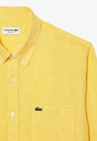 Camisa amarilla de algodón, hecha de lino, con un bolsillo en el pecho y un pequeño logo de cocodrilo bordado en verde. Corte regular, tela texturizada.
