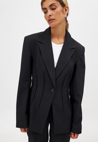 Black tailored blazer with structured shoulders, single button closure, and vertical seam details, made from smooth fabric. Plain white top underneath.