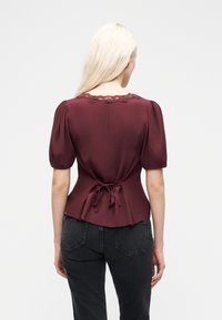 Γυναίκα με μακριά ξανθά μαλλιά, φορώντας εφαρμοστή μπλούζα μπορντό με φουσκωτά μανίκια και δαντελωτή  επένδυση, δεμένη στο πίσω μέρος, συνδυασμένη με μαύρο τζιν.