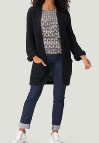 Schwarze Strickjacke mit offenem Vorderteil, strukturierten Ärmeln und zwei Taschen. Kombiniert mit einem gemusterten Oberteil, dunklen Jeans und weißen Sneakers.