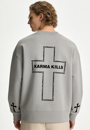 Persoon met krullend haar draagt een grijze trui met een zwart kruis gemaakt van herhaalde "KARMA KILLS" tekst en kleinere kruisen op beide mouwen.