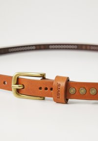 Ceinture en cuir brun avec une boucle en laiton, dotée de clous décoratifs sur une sangle beige, marquée "Levi's". Fini texturé, avec plusieurs trous pour l'ajustement.