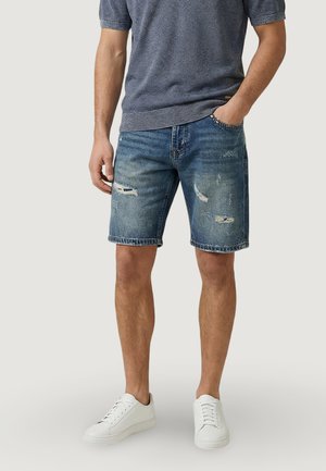 Mężczyzna w wyblakłych niebieskich, przetartych szortach z denimu, białych trampkach i szarej, teksturowanej koszulce z krótkim rękawem, stojący z jedną ręką w kieszeni.