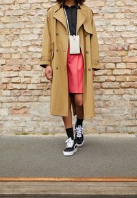 Persona con un trench coat beige, maglietta nera, gonna rossa, calze nere e sneakers bianche e nere in piedi contro un muro di mattoni.