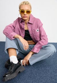 Veste en denim rose avec une poche poitrine, débardeur noir, pantalon large gris clair, et chaussures à plateforme noires. Lunettes de soleil rondes jaunes et boucles d'oreilles créoles.