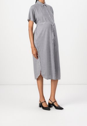 Nainen, joka pukeutuu mustavalkoiseen pystyraitaan, midi-pituisen paitamekon lyhyillä hihoilla ja mustiin Mary Jane -kengillä, joissa on paksu korko.