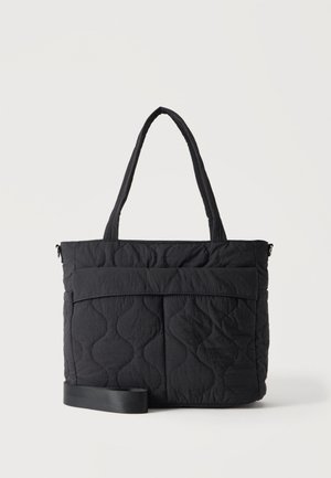 Sort quiltet tote taske med to håndtag og en justerbar skulderrem, med en front flap lomme på en ensfarvet baggrund.