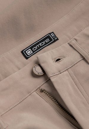 Pantalons beiges avec une texture lisse, présentant une fermeture éclair et un bouton en métal rond. L'étiquette de la marque "ombre" est cousue en noir et blanc.