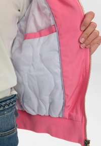 Interno di una giacca bomber rosa che mostra una fodera trapuntata bianca con una tasca rosa, una zip dorata e un orlo a costine. Texture liscia con dettagli sottili.