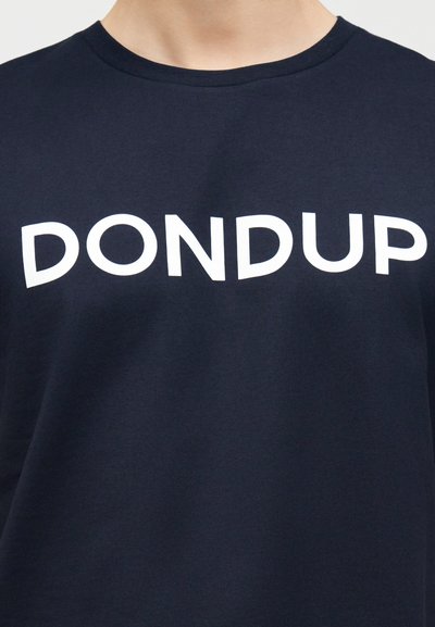 Άτομο που φοράει σκούρο μπλε μπλουζάκι με στρογγυλή λαιμόκοψη και μεγάλο λευκό κείμενο "DONDUP" τυπωμένο στο στήθος.