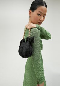 Bolso negro texturizado con acentos anudados y una cadena dorada, sostenido por una persona con un vestido verde estampado.