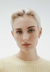 Suéter de punto acanalado de manga corta en amarillo pálido. La modelo tiene el pelo corto, rubio claro y lleva pequeños pendientes de aro dorados. Fondo neutro.