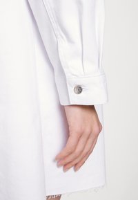 Punho de camisa de algodão branco com um botão de pressão prateado, apresentando uma textura lisa e um ligeiro desfiar na bainha. Mão repousando suavemente sobre o tecido.