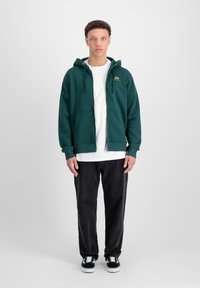 Felpa verde con zip e tasche a marsupio, indossata su una t-shirt bianca. Pantaloni neri a vestibilità ampia e sneakers nere completano il look.