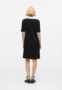 Černé šaty s krátkým rukávem a bílým límečkem, hladká textura, ke kolenům, volný střih; nošeny s černými střevíčky se cvočky.