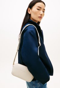 Γυναίκα με μακριά σκούρα μαλλιά που φοράει ένα ναυτικό μπλε σακάκι και τζιν, κρατώντας μια μικρή μπεζ τσάντα Tommy Hilfiger στον ώμο.