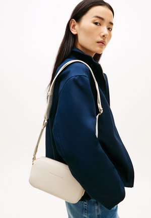 Vrouw met lang donker haar die een marineblauw jack en spijkerbroek draagt, met een kleine beige Tommy Hilfiger schoudertas.