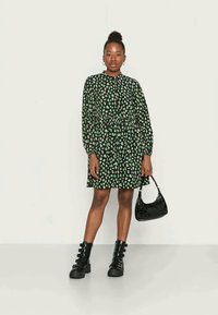 Vestuário preto com padrão de folhas verdes, mangas longas e cintura ajustada, combinado com uma bolsa preta em couro sintético e botas pretas robustas.