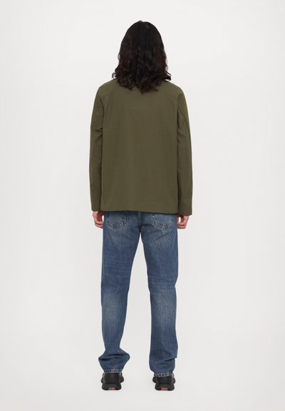 Veste vert olive à coupe droite, associée à un jean en denim bleu. La veste présente un design simple et une texture uniforme.