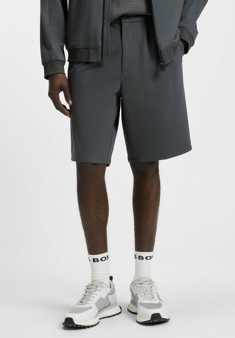 Pantaloni corti sportivi grigio scuro in tessuto liscio, con vita strutturata e fronte piatto. Abbinati a calze bianche e scarpe da ginnastica grigie.