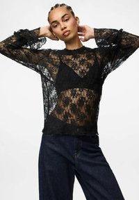 Crna prozirna bluzica od čipke s cvjetnim uzorcima, okruglim izrezom, dugim rukavima s volanastim manžetama, prikazana preko crnog grudnjaka i traperica.