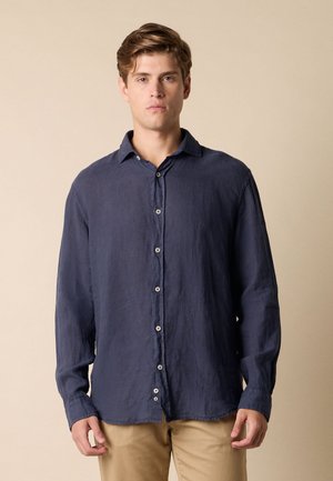 Junger Mann mit kurzen hellbraunen Haaren, der ein langärmeliges marineblaues Hemd mit Knöpfen und beigen Hosen trägt und vor einem beigen Hintergrund steht.