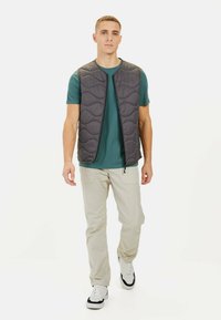Polstret gråt vest med rund halsudskæring, frontlynlås og quiltet design, båret over en grøn skjorte og parret med lysebeige bukser og sneakers.