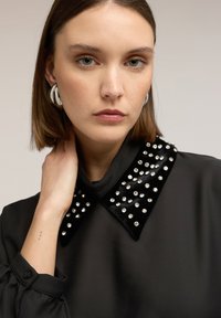 Blusa negra con un cuello de terciopelo adornado con tachuelas plateadas. Mangas largas con puños fruncidos y una textura de tela lisa.