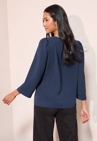 Marineblauwe blouse met driekwartmouwen en een gestructureerde stof, gecombineerd met zwarte broek met wijd uitlopende pijpen, weergegeven vanuit een zijaanzicht.