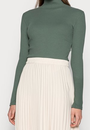 Kvinde iført en tætsiddende, ribstrikket grøn rullekrave og et højtaljet, hvidt plisseret skørt, stående med armene afslappet langs siderne.