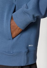 Sudadera azul con puños y dobladillo acanalados, con un bolsillo que tiene una mano dentro. Logo "DRYCELL" en blanco cerca del dobladillo, textura de tela suave.