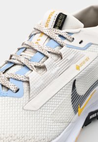 Les chaussures Nike Pegasus Trail 5 présentent une tige en mesh blanche et bleu clair avec des accents noirs et jaunes, des surfaces texturées et une étiquette Gore-Tex.