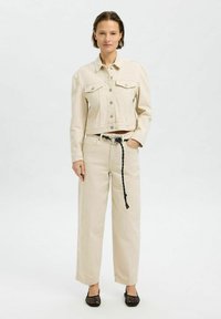 Beige denim cropped jas met twee borstzakken, gecombineerd met beige broek met brede pijpen, voorzien van een zwarte gevlochten riem en zwarte mesh schoenen.
