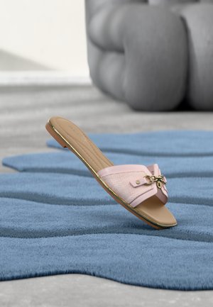 Różowa płaska sandałka typu slide z ozdobą ze złotego łańcucha, spoczywająca na niebieskim, teksturowanym dywanie, na tle rozmytej szarej pikowanej sofy.