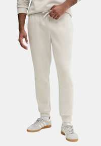 Jogger beige chiaro con vita elasticizzata e cordoncino, caviglie affusolate e superficie liscia, abbinati a sneakers grigie con tre strisce bianche.