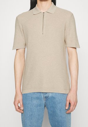 Homme portant un polo en maille beige à manches courtes avec une fermeture éclair sur le devant et un jean bleu clair.