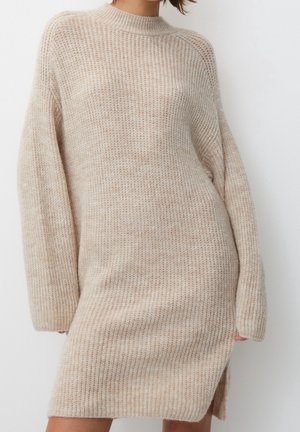Kvinde iført en beige, langærmet, strikket sweaterkjole med en sideslids, stående mod en ensfarvet hvid baggrund.
